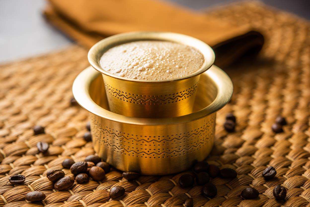 Frothy coffee in a traditional brass tumbler and davara, surrounded by scattered coffee beans.