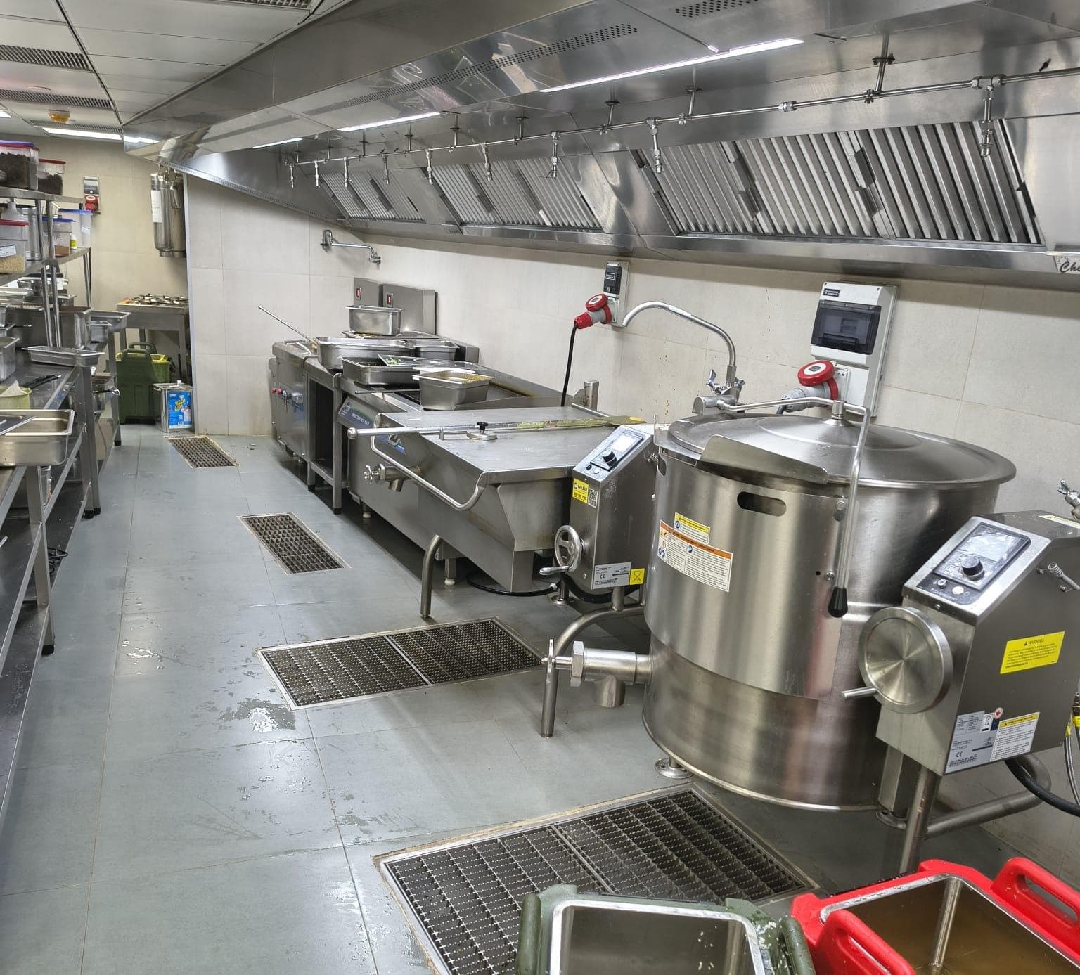 Professional kitchen featuring large stainless steel cooking kettles, tilting skillets, and industrial ventilation.