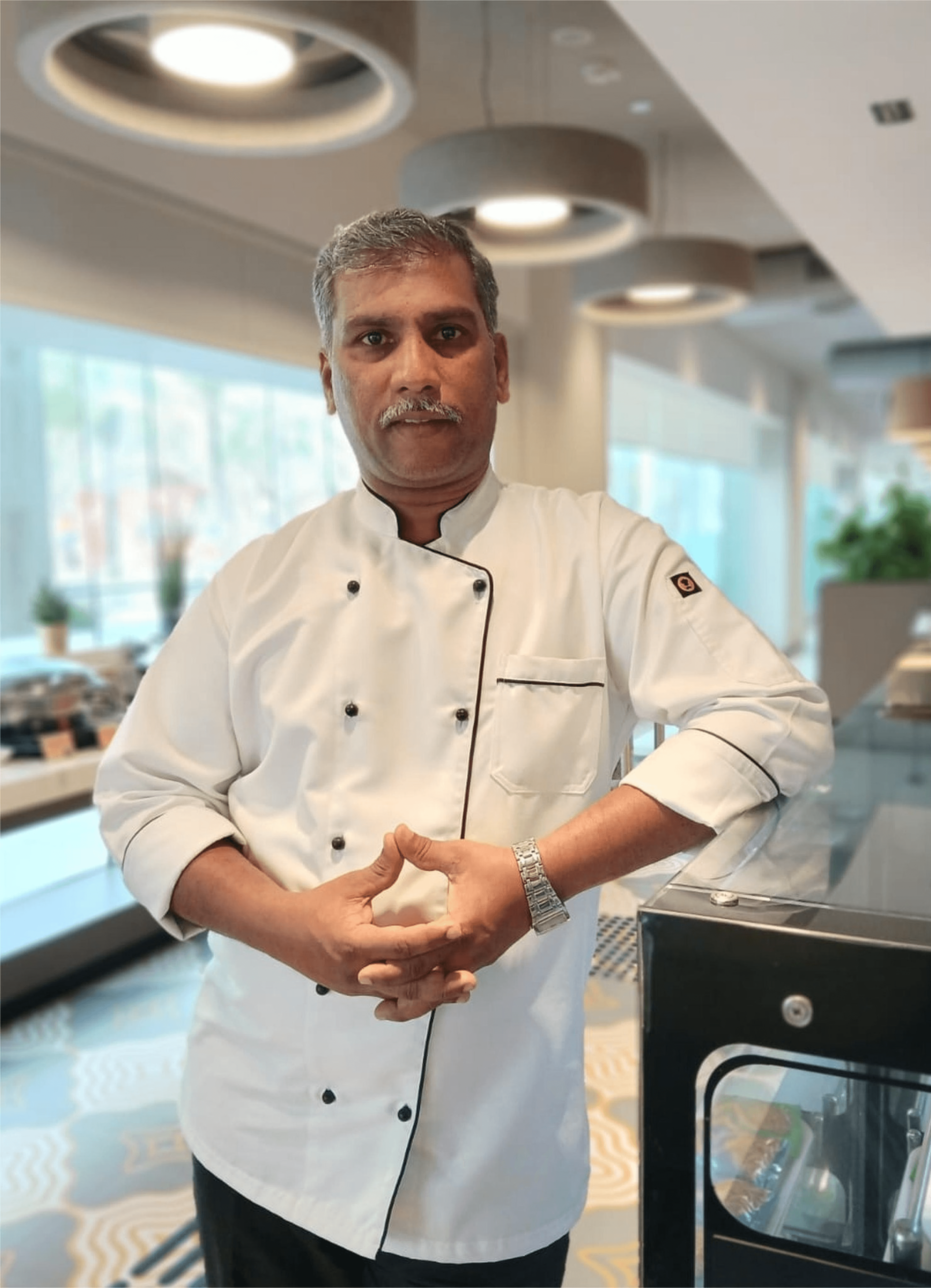 Professional chef in a white double-breasted coat posing in a bright, contemporary dining area.