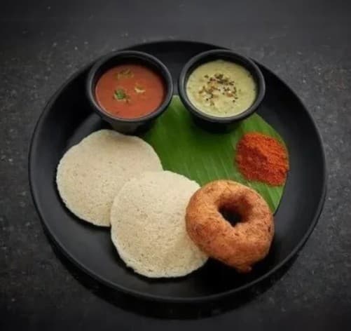 Two idlis and a medu vada served with chutneys and podi on a banana leaf.